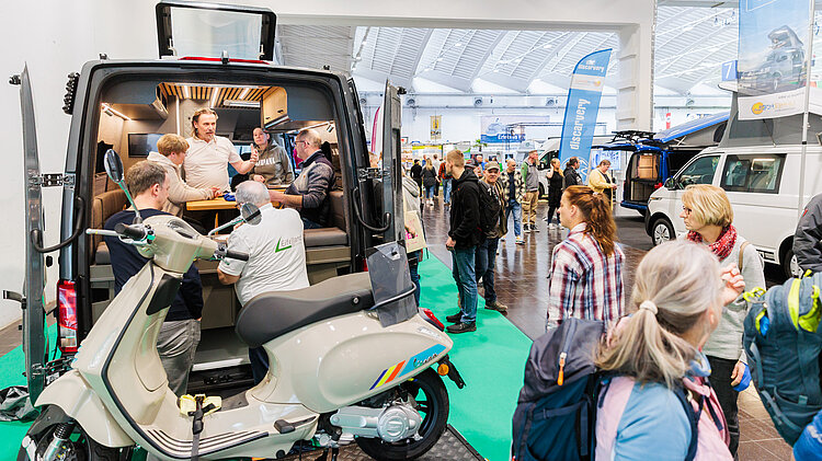 Besucherinnen und Besucher stehen in einer Messehalle vor einem ausgebauten Camper-Van und informieren sich über Vanlife, Ausbauten und Zubehör auf der Reise + Camping 2026.