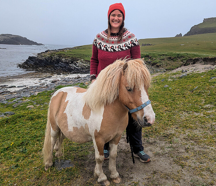 Frau in Wollpullover steht an der Küste der Shetlandinseln neben einem Shetlandpony