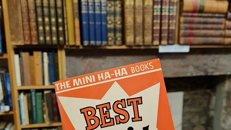 Hand hält ein kleines Buch mit dem Titel „Best Scottish Jokes“ vor hohen Bücherregalen in einem Antiquariat
