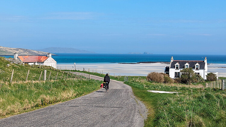 Radfahrerin auf schmaler Straße auf den Äußeren Hebriden mit Blick auf Meer, Strand und einzelne Häuser